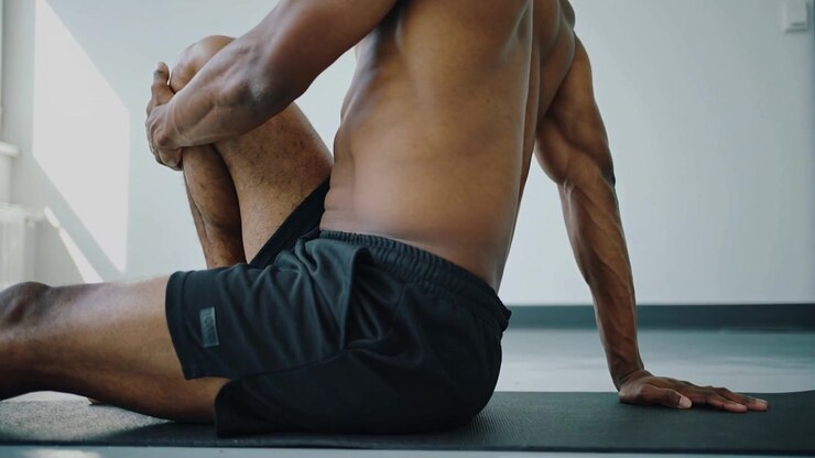 Sistemandosi sul tappetino, un uomo a torso nudo in pantaloncini neri porta il ginocchio sinistro al petto facendo stretching in studio