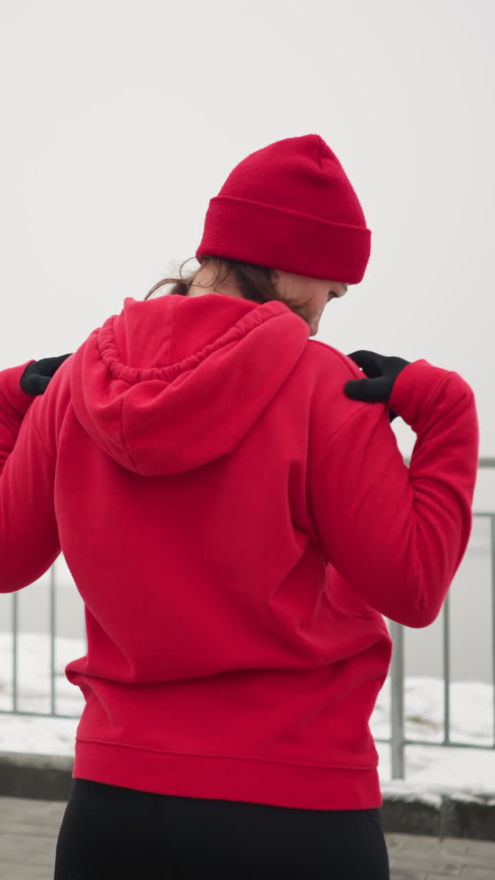 back view of coach in red hoodie outdoors performing neck stretches with hands on shoulders, turning head left and right in serene winter setting with snow-covered ground and iron railing