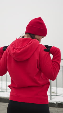 back view of coach in red hoodie outdoors performing neck stretches with hands on shoulders, turning head left and right in serene winter setting with snow-covered ground and iron railing