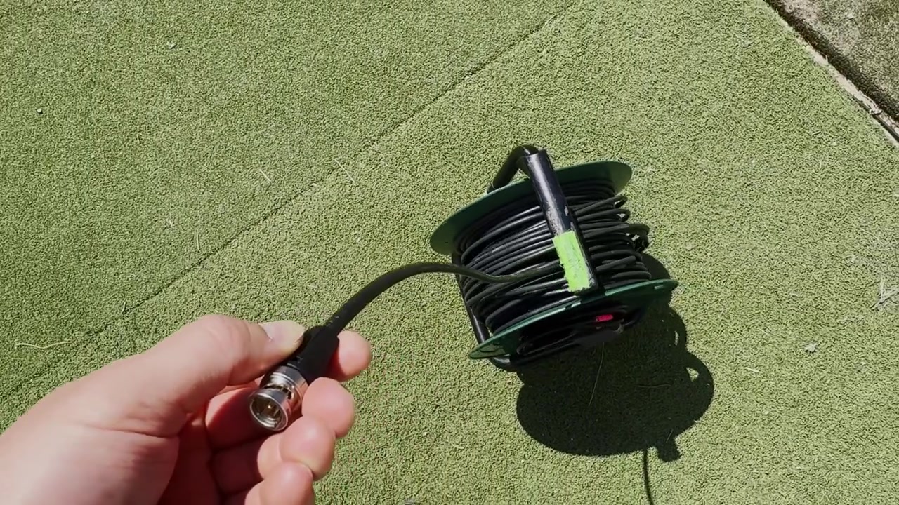 Hand holding an SDI BNC video cable connector beside a coiled cable reel on artificial turf, showing broadcast production wiring and outdoor setup detail.