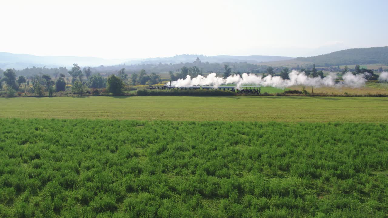 A panoramic aerial shot captures a historic steam train on a peaceful journey. The locomotive puffs billows of white steam as it crosses lush green fields in the vast countryside