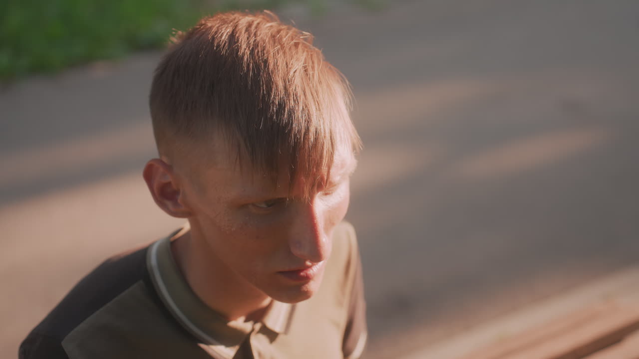 Young White Man On Bench In Golden Hour Light, Head Down And Thoughtful With Tense Jaw And Distant Eyes Warm Shadows, Urban Park Walkway, Reflective Mood, Subtle Signs Of Worry And Mental Strain