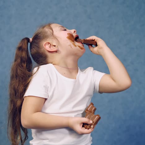 cute girl licking bars of chocolate indoors. happy child eating delicious sweets and becomes dirty. little kid is smeared with chocolate.