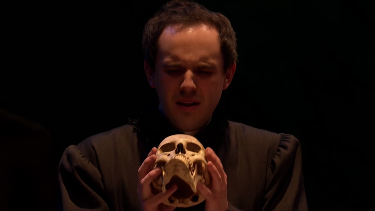 Attore caucasico che tiene in mano un teschio in un teatro buio. Atmosfera contemplativa con chiaroscuro a lume di candela, indossa una tunica nera.