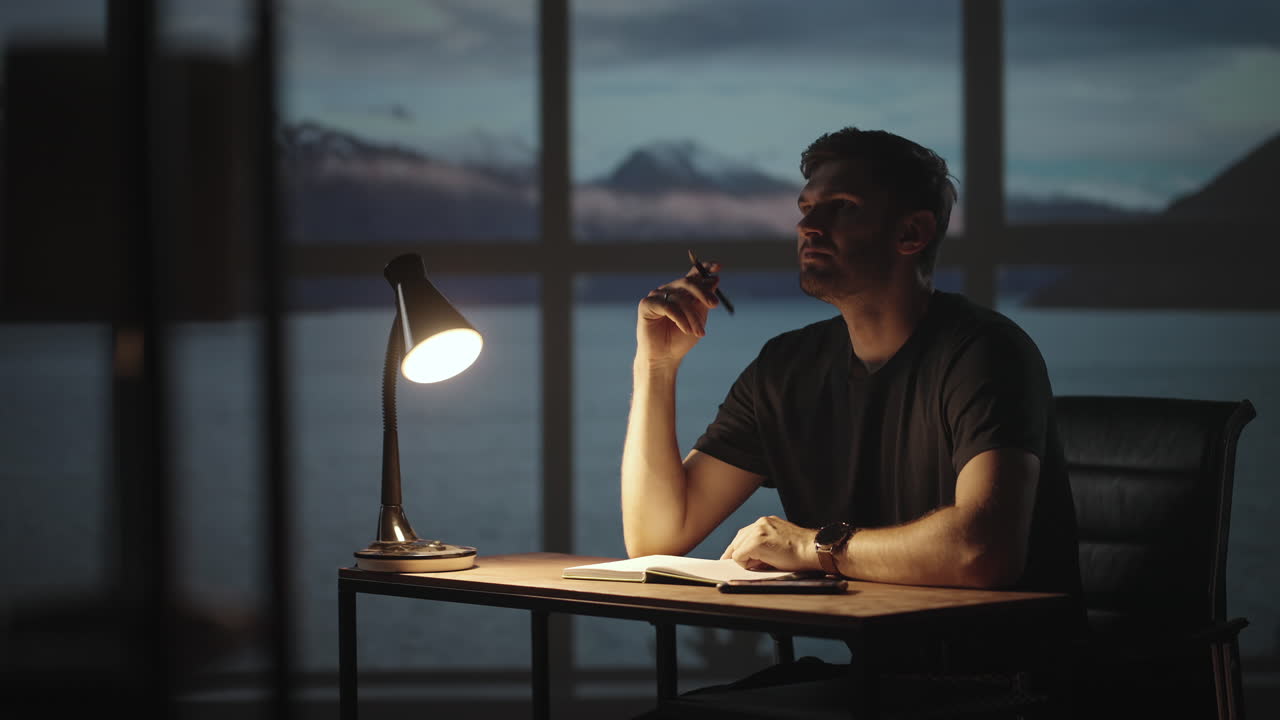 The silhouette of a thoughtful man sitting at a table against the background of a window with oceans and the sea. A restless man sitting at a table with a desk lamp