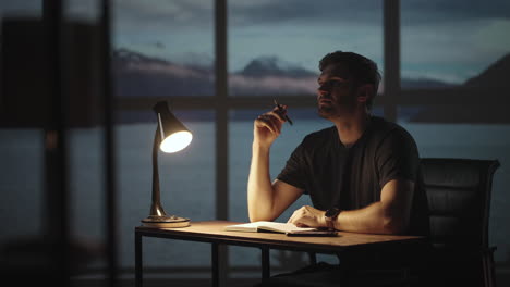 the silhouette of a thoughtful man sitting at a table against the background of a window with oceans and the sea. a restless man sitting at a table with a desk lamp