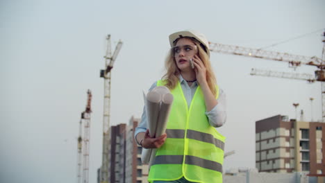 mujer constructor hablando por teléfono con el contratista con dibujos en sus manos en el fondo de los edificios en construcción y grúas