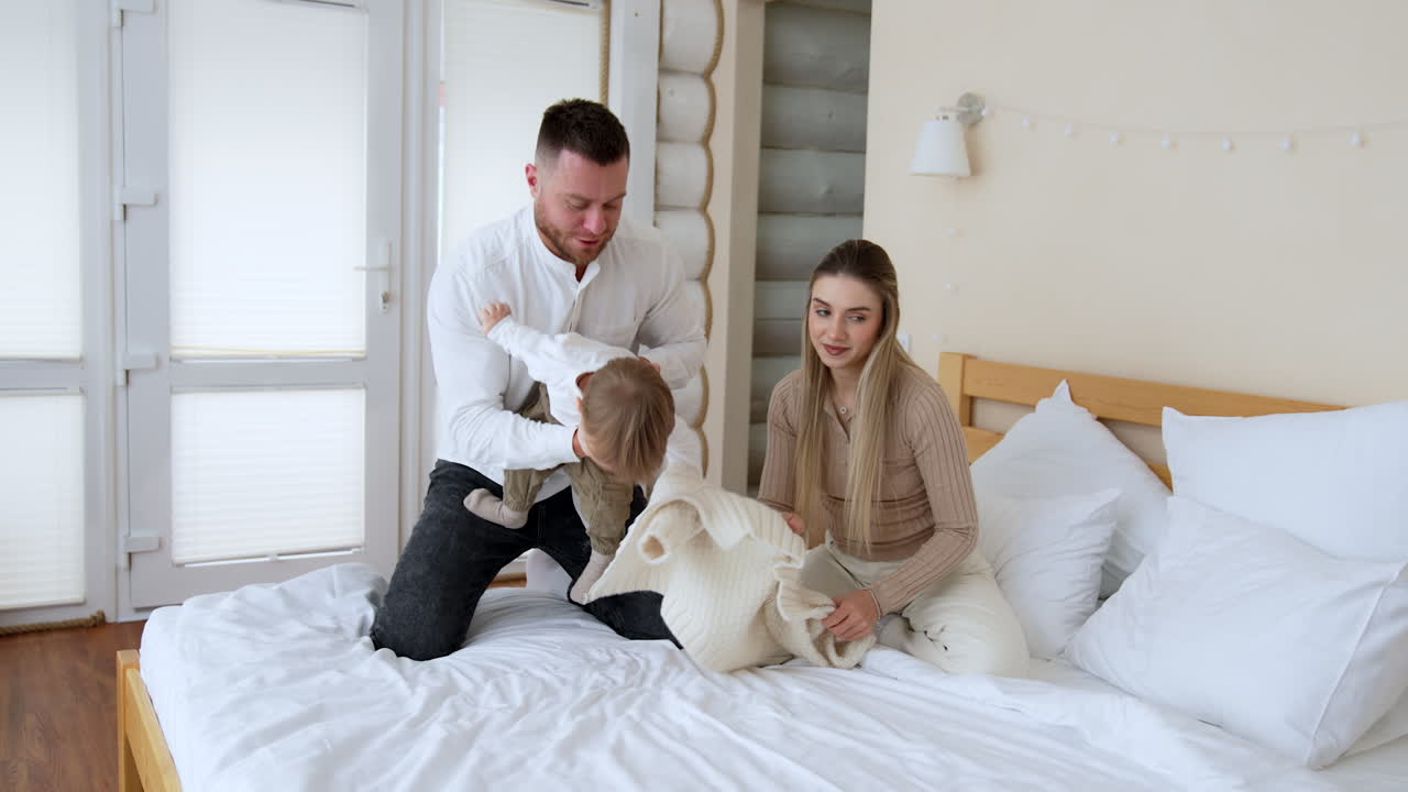 Father tumbles his baby boy putting him on the bed. Parents take off kid's warm sweater. Beautiful family portrait.
