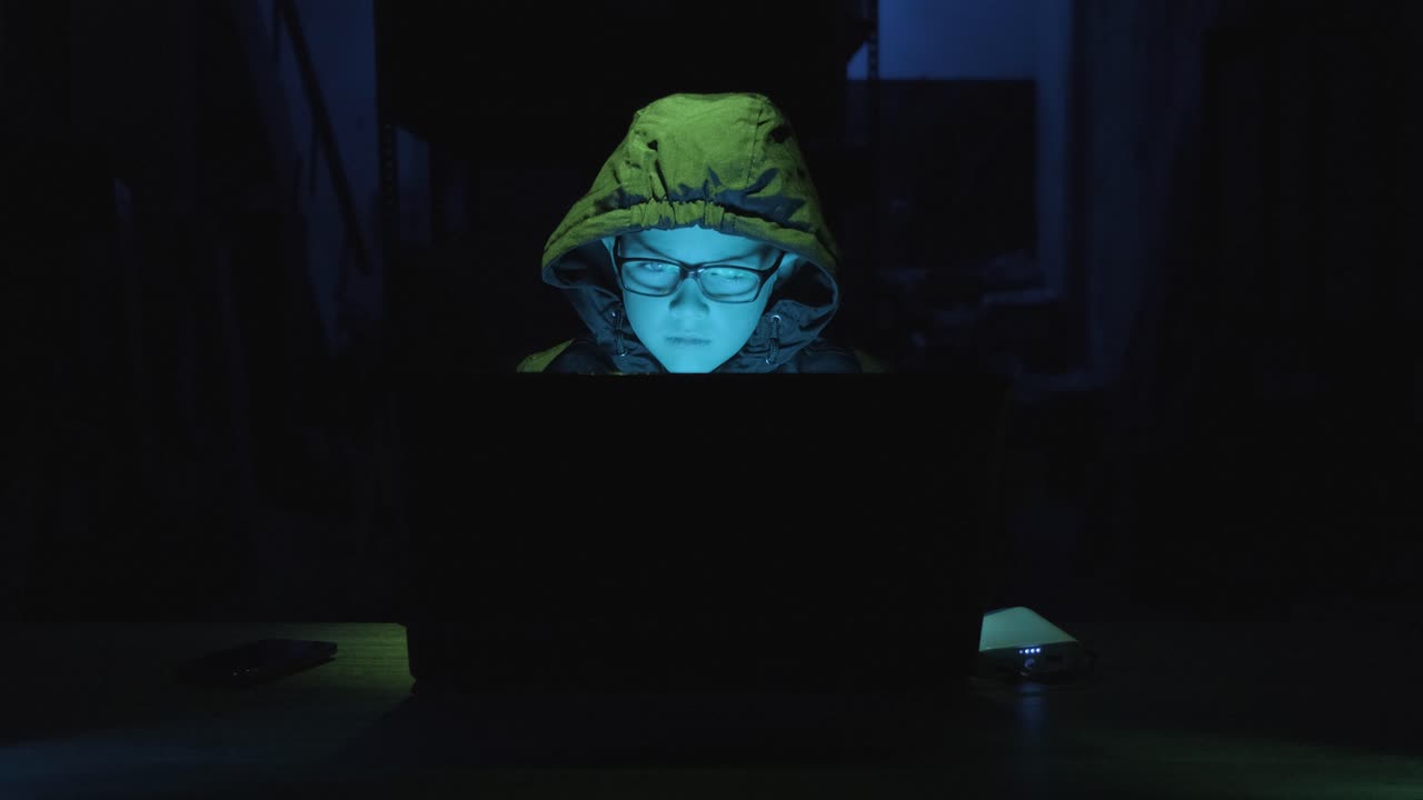 Young Boy Hacker In Hoodie And Eyeglasses Using Laptop In Dark Room. medium shot, zoom-in