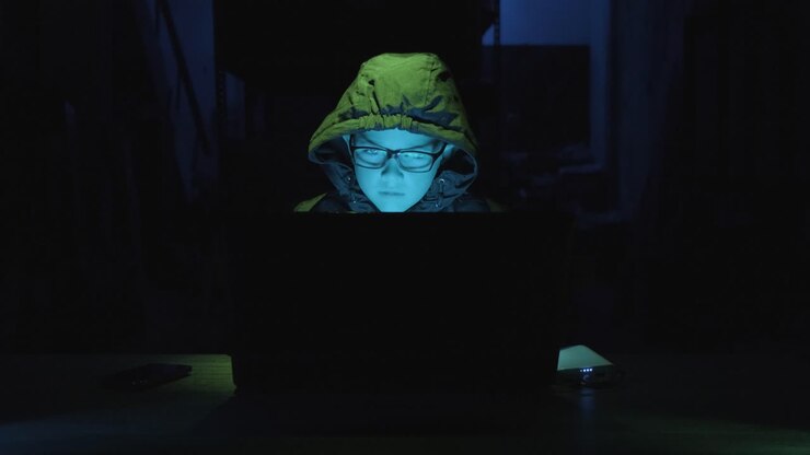 Giovane hacker con felpa con cappuccio e occhiali che usa un laptop in una stanza buia. Inquadratura media, zoom in.