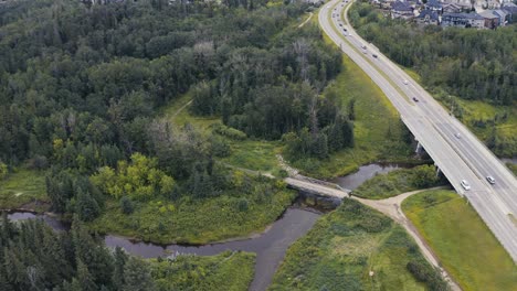 Aerial-fly-over-highway-freeway-from-a-forest-park-to-the-light-traffic-town-road-next-to-a-cookiecut-luxury-community-by-a-romantic-breathtaking-river-with-a-vintage-bridge-where-weddings-take-place