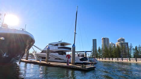 a large luxury yacht slowly reverses into a marina dock under bright sunlight, with modern city buildings and calm water in the background