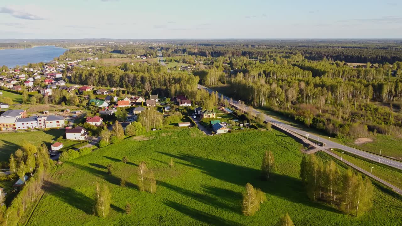 Flying over Katlakalns district in springtime, the camera shows houses, Daugmale road, fields with tall shadows, and the Daugava River in the distance under clear sky, slow motion