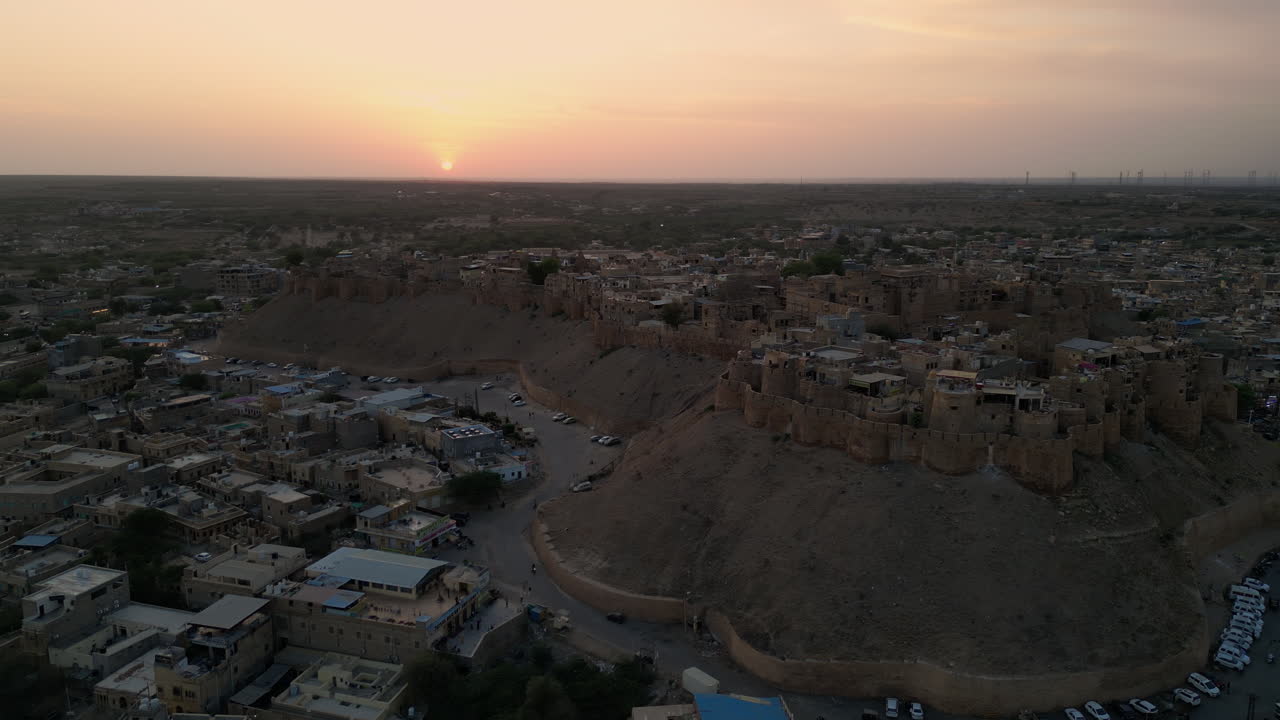 una toma de órbita de un dron de 4k del fuerte de jaisalmer durante la puesta del sol con el sol visible en el marco, arrojando un brillo cálido sobre el fuerte de rajasthan, india