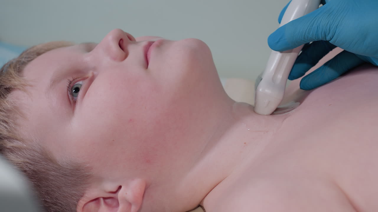 Close up of young boy lying calmly while female medical professional in blue gloves performs throat ultrasound examination using transducer with gel during clinical diagnostic procedure in a hospital