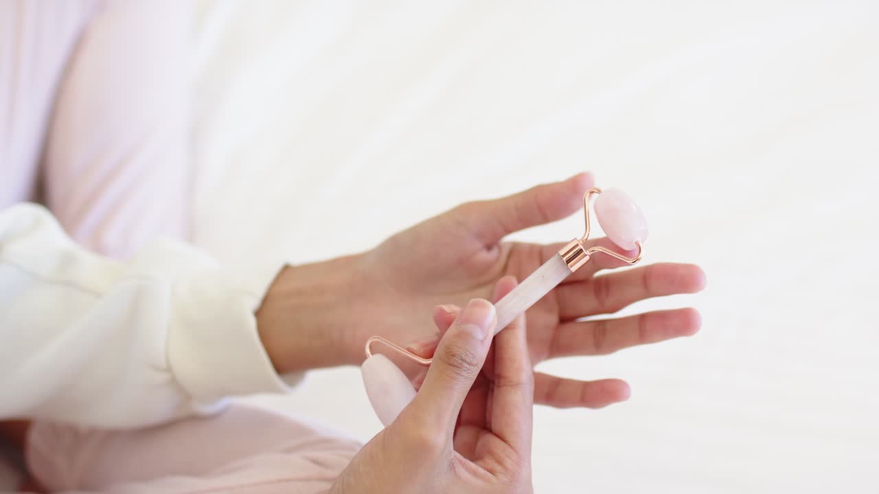 Le mani iniziano sostenendo delicatamente e poi ruotando un rullo facciale di quarzo rosa sul letto, assicurandosi che scorra liberamente.