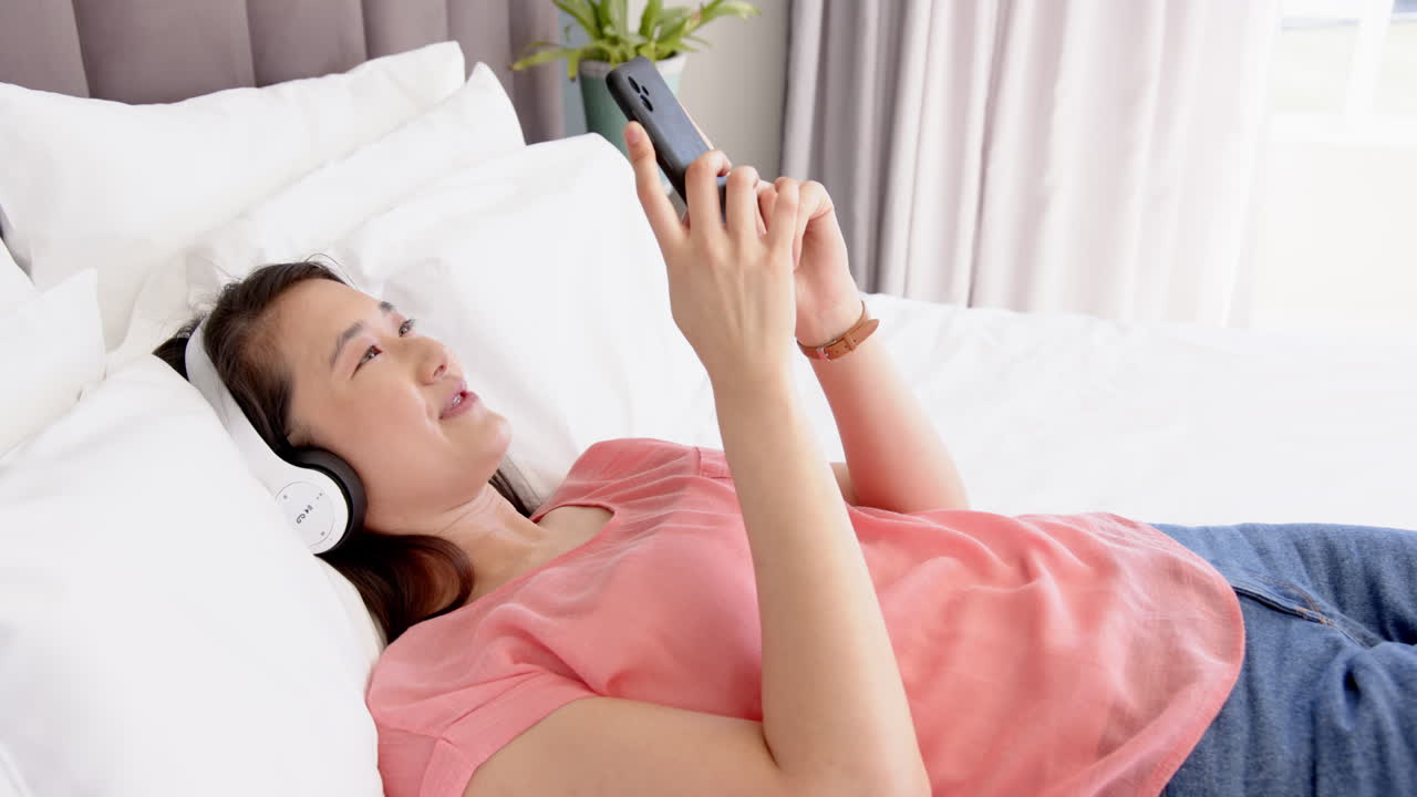 Lying on bed, woman smiling while using smartphone and wearing headphones