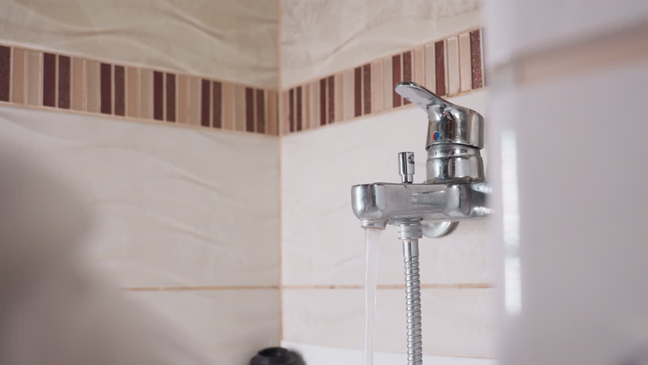 Lower view shows woman hand near chrome mixer as shower water rushes from spout, fingers reach lever to turn flow off, rippled tiles reflect light, hose dangling, calm bathroom routine