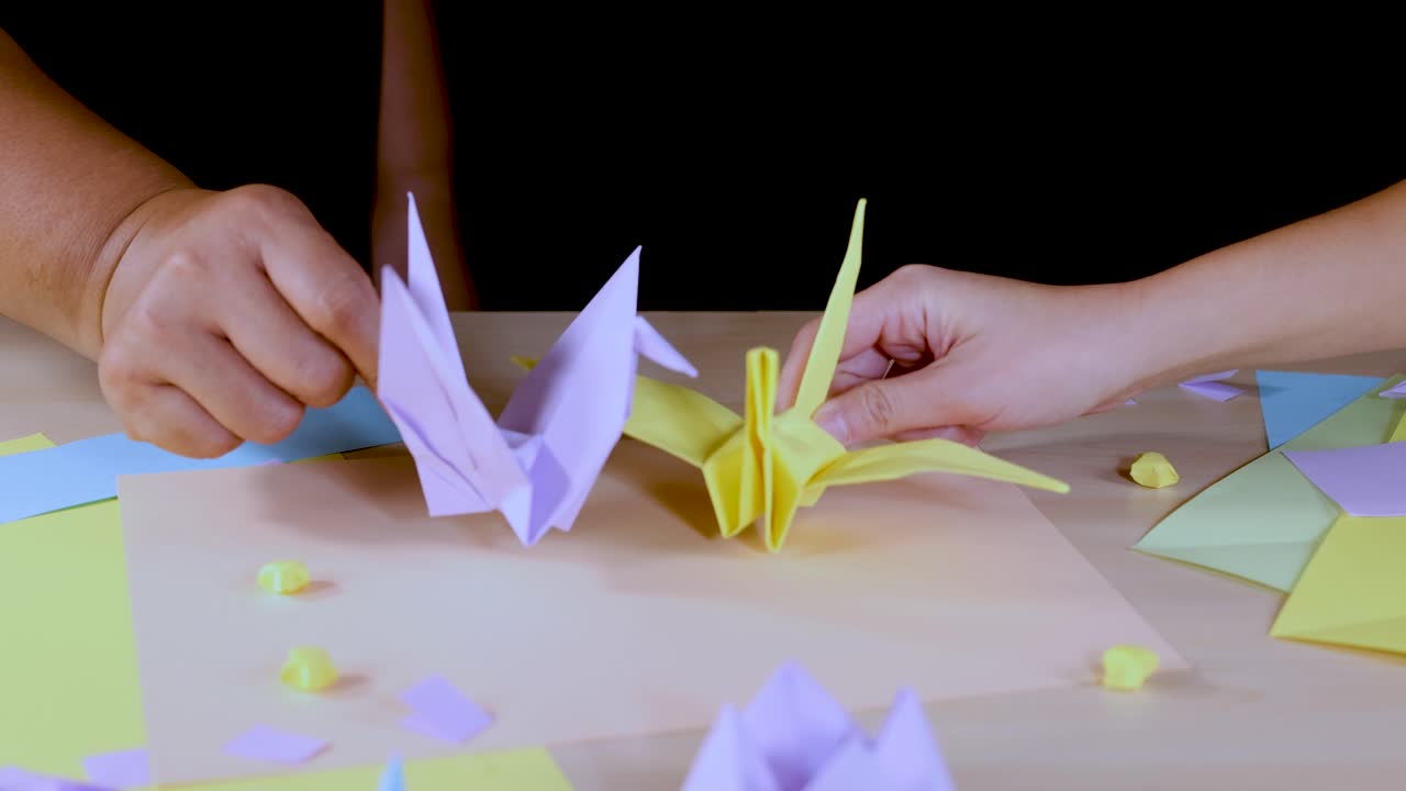 Adult hands arrange pastel origami cranes on a well-lit table with colored paper sheets