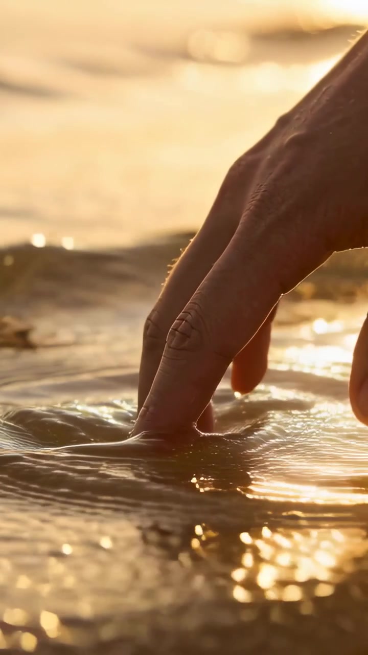 Vertical video: Descending hand touching shallow shore water, creating ripples and sunlit glints