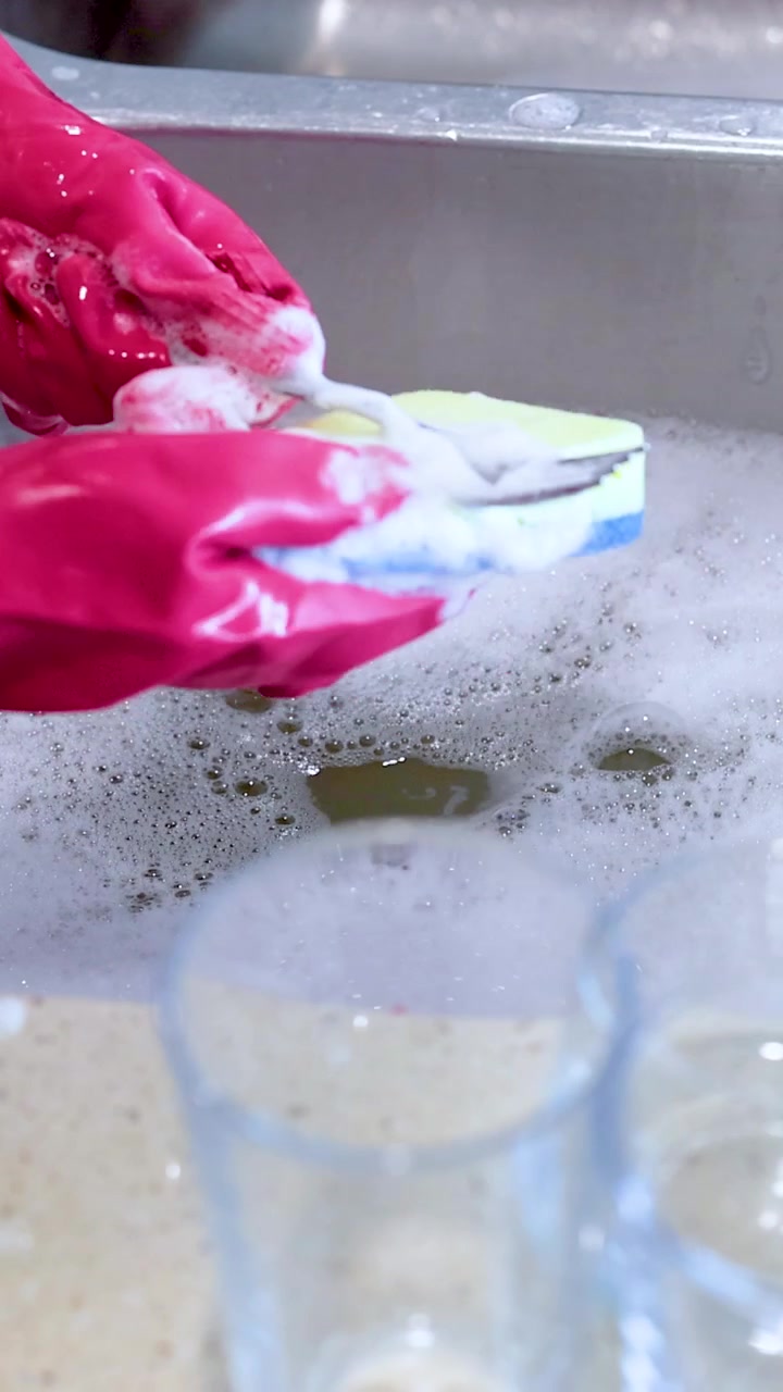 Hands in pink gloves clean a fork with a sponge over a soapy sink. Bright kitchen lighting