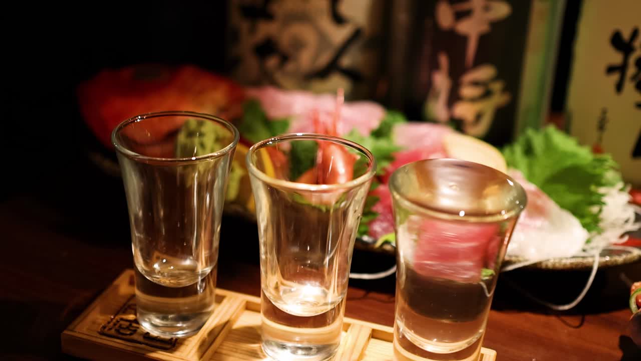 Close-up of sake glasses with sushi and garnish on a wooden table in ambient lighting.