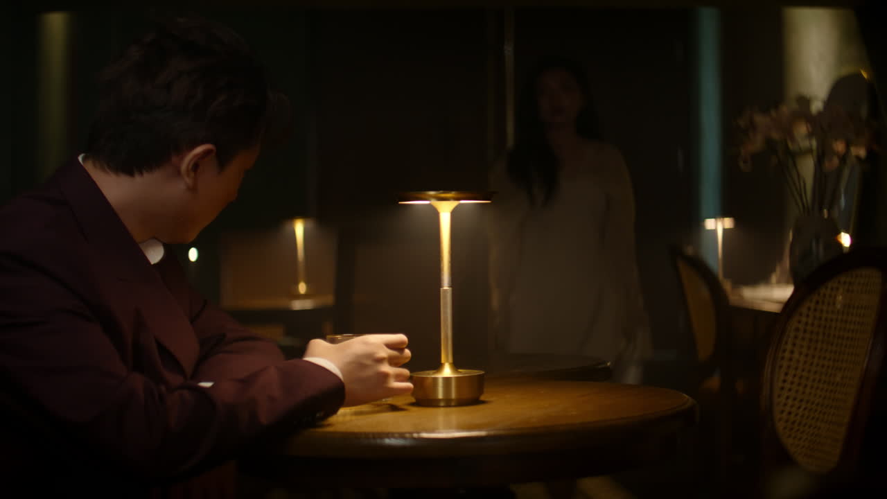 A man in a suit sitting alone at a table, lost in thought, while a woman walks by