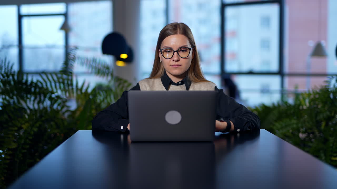 Lady in glasses opens laptop, prints something and closes it. Long-haired woman puts off glasses and lays her head on hands. Blurred backdrop.
