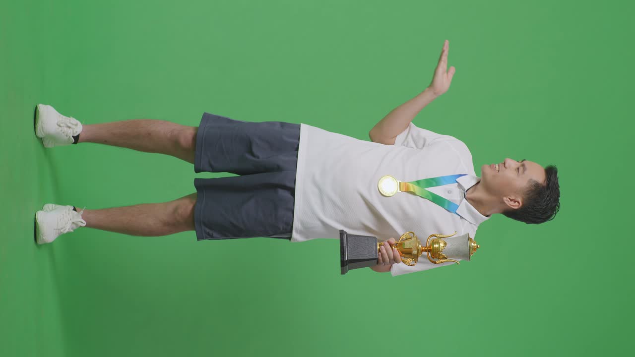 황금 메달을 가진 아시아 남자의 몸이 가득 차 있으며, 손에 금 트로피를 들고 미소 짓고, 스튜디오의 녹색 화면 배경에 을 가리키고 있습니다.