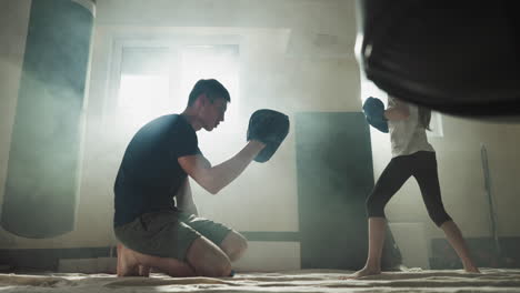 el niño patea la almohadilla con el apoyo del entrenador en el gimnasio de combate. el entrenador de kickboxing enseña a la niña descalza el ataque con la pierna en la escuela de deportes en cámara lenta. lección de autodefensa