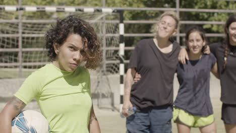 serious female football player holding a soccer ball and looking at the camera while standing on the field with her team