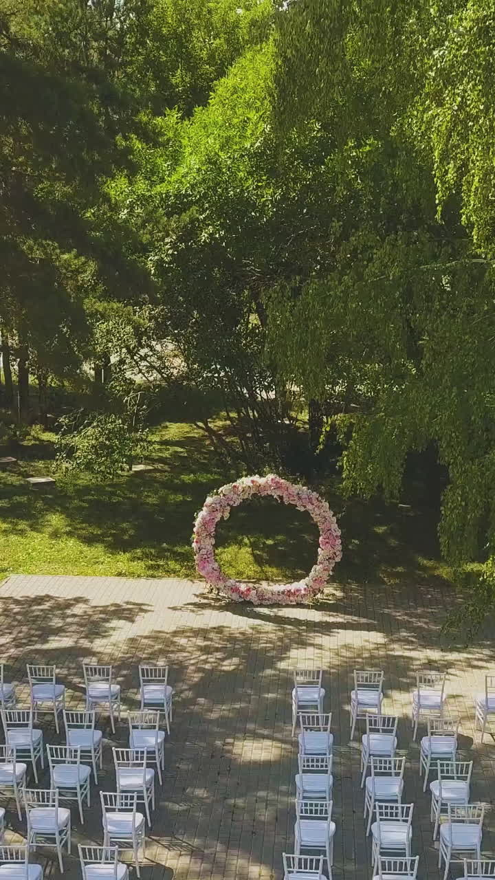 wedding venue decorated with pink flower garland and chairs in picturesque green park on autumn day aerial view