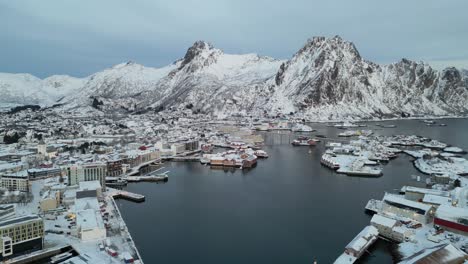 svinøya village surrounded by snow-covered mountains and a calm harbor , aerial view