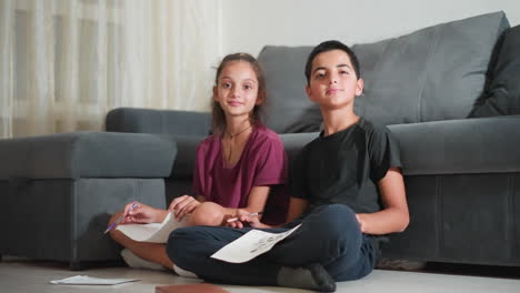 cute siblings seated near couch holding pieces of paper smiling at camera in well-lit cozy living room showing joy of creative time together with sketchpads bonding over shared drawing