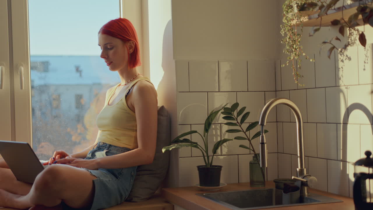 hermosa chica sentada en el alféizar de la ventana iluminada por el sol en casa y usando una computadora portátil