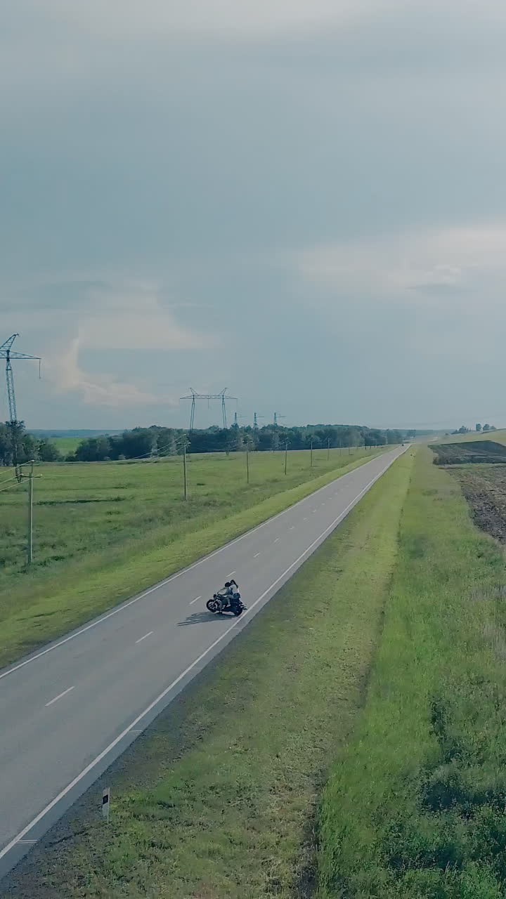 bird eye view couple on motorcycle lit by summer sunlight turns back and drives near pictorial green grass