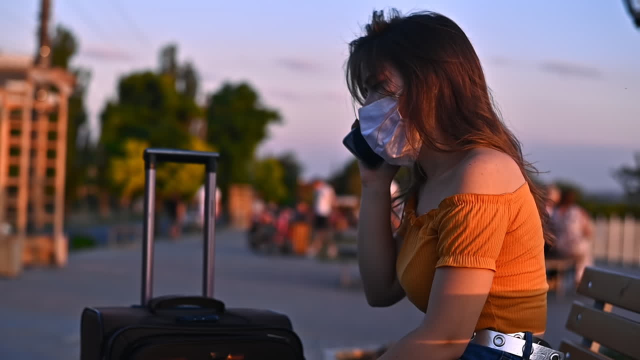 Young woman talking on the phone while resting on the bench at sunset. Wearing protective medical mask and carrying a luggage. Corona Virus idea. Safe travel during the pandemic. Slow motion