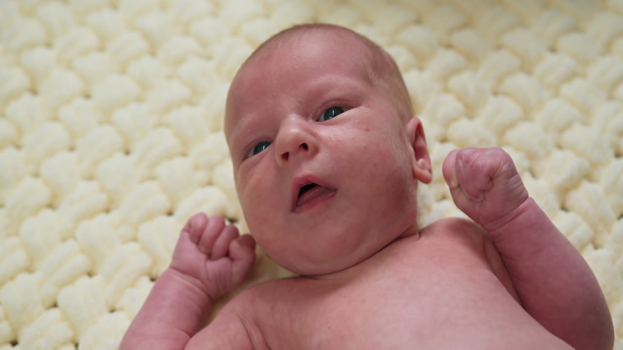 Nahaufnahme eines neugeborenen Babys mit geballten Fäusten, das auf einer cremefarbenen Decke liegt und die Augen geöffnet hat
