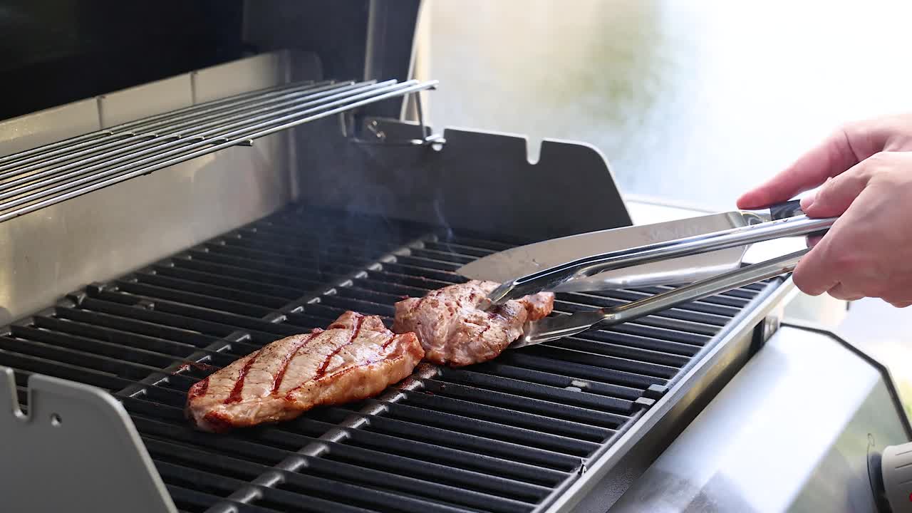 Hands flipping a grilled beef steak on a barbecue grill. Bright outdoor lighting enhances the cooking process