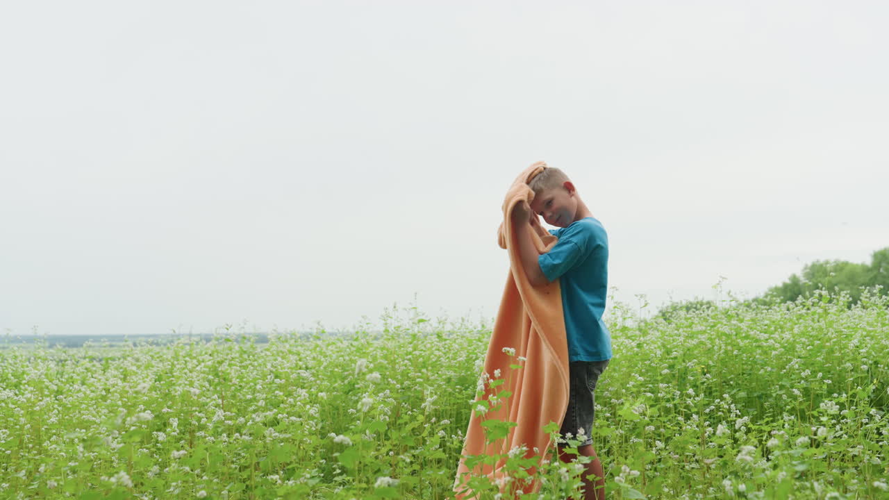 Young boy surveys cloth amidst blooming flowers, Young boy observes fabric among wildflowers beneath cloudy sky, Solitary child inspects orange cloth surrounded by summer greenery under grey clouds