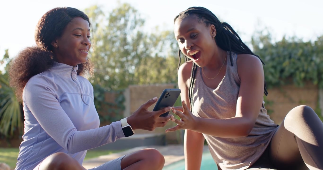Two adult women sitting on poolside mat in activewear offering smartphone, reaching to view content