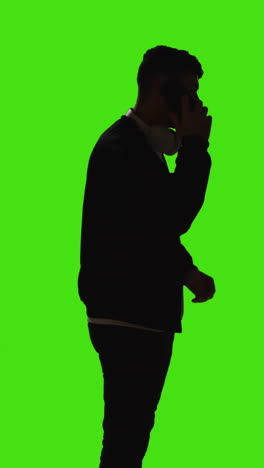 Vertical-Video-Silhouette-Of-Man-With-Wireless-Headphones-Answering-Call-On-Mobile-Phone-Against-Green-Screen