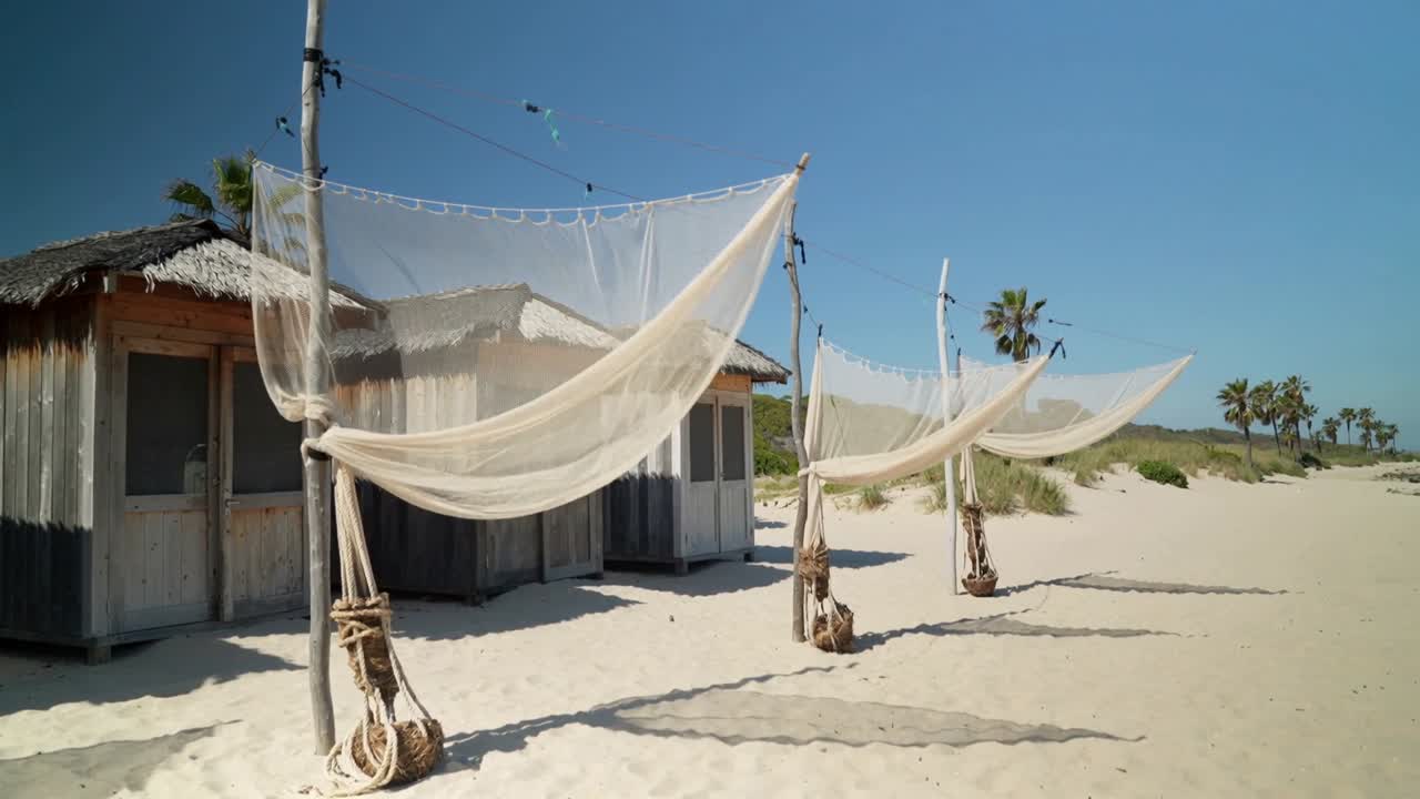 Fila ondeggiante di amache in rete vicino a una baracca logora, che reagisce alla leggera brezza marina su una spiaggia sabbiosa.