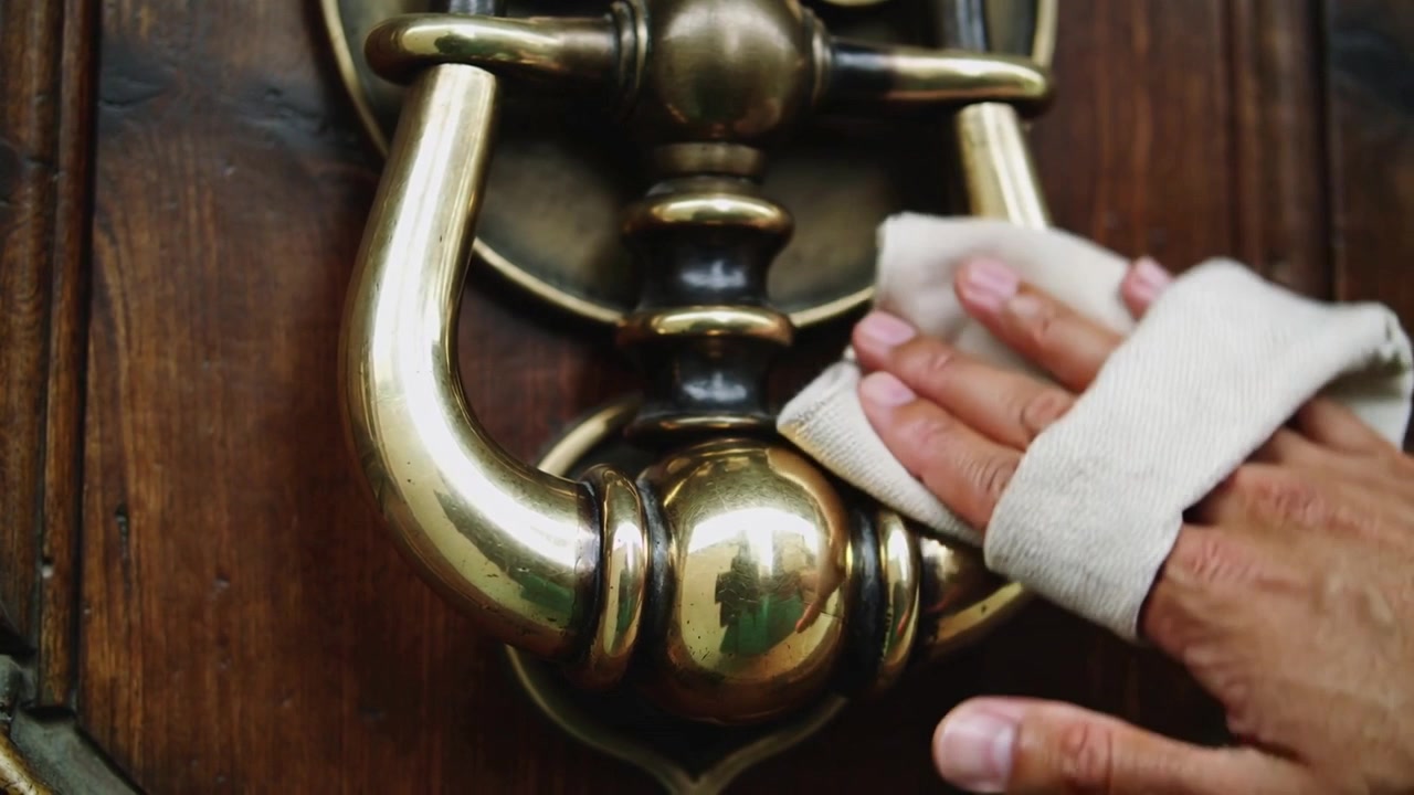 Mani che posizionano una cinghia di stoffa bianca sul battente e lucidano l'ottone intarsiato della porta d'ingresso per ripristinare la lucentezza.