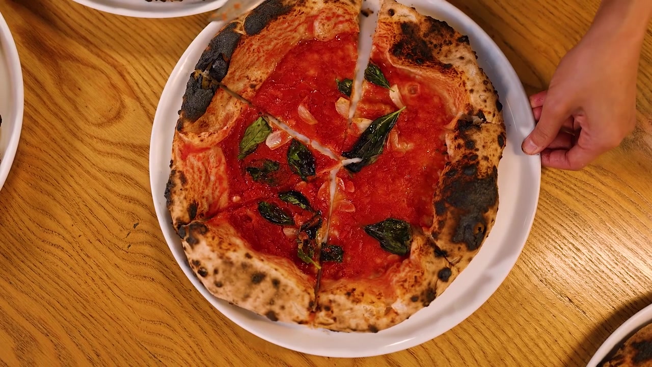 Ripresa dall'alto di mani che affettano una pizza a crosta sottile con salsa di pomodoro e basilico