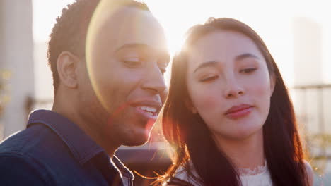 Couple-Sitting-On-Rooftop-Terrace-Against-Flaring-Sun-With-City-Skyline-In-Background