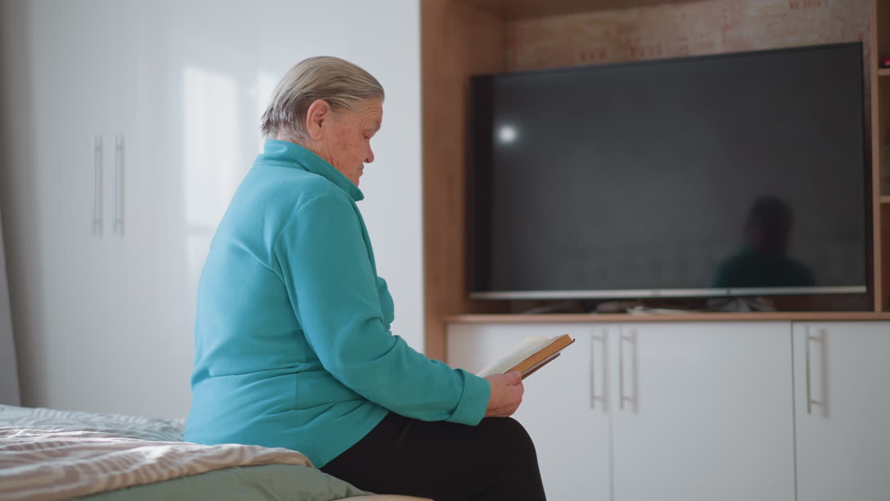 Elderly woman in teal green sweater sitting on bed reading a book, soft natural light from window, TV in background, peaceful indoor setting with minimalistic decor, quiet and reflective moment
