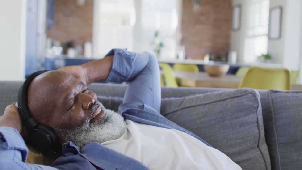 Relaxed senior african american man in living room lying on sofa, wearing headphones