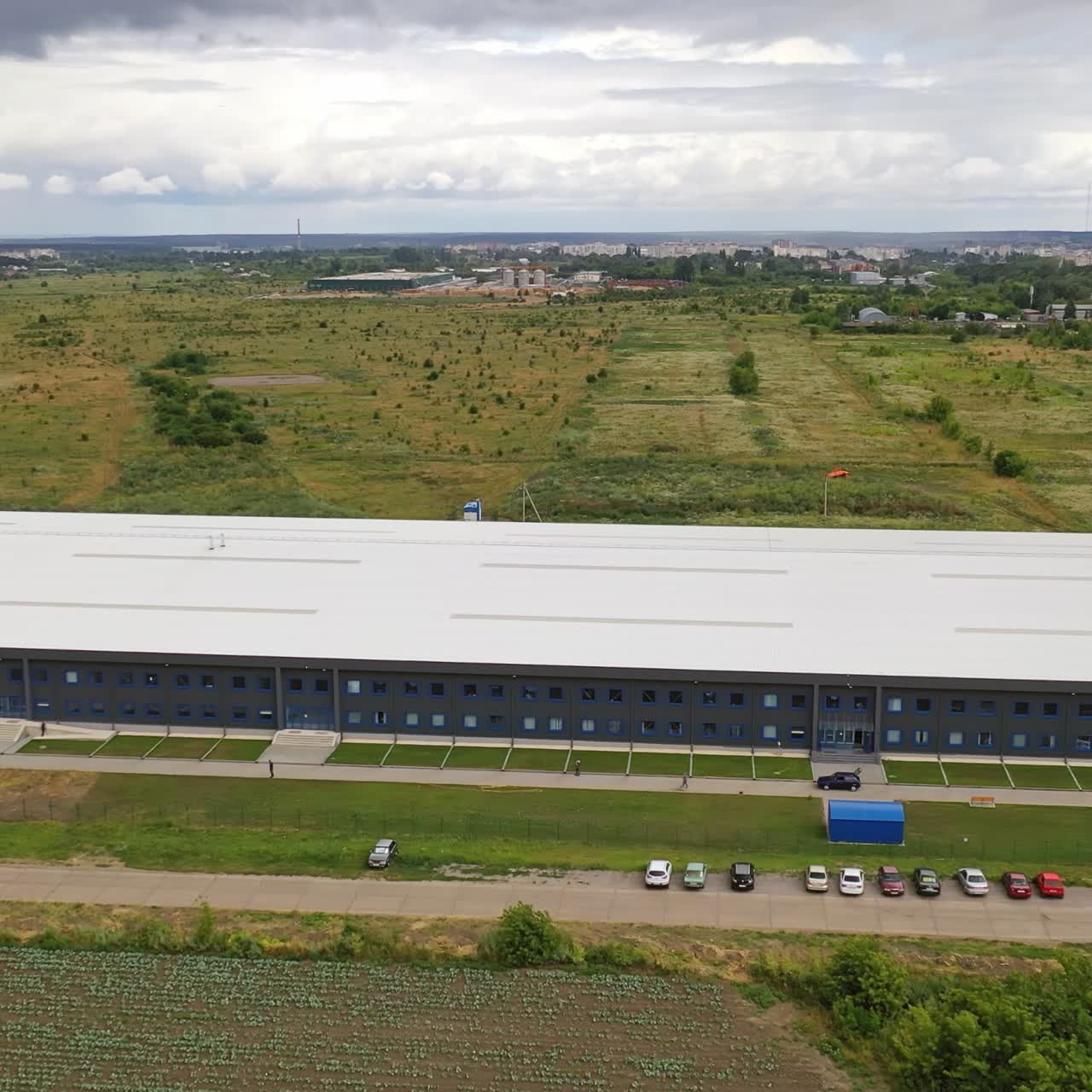 Aerial panorama of a modern plant. Exterior of a large commercial warehouse situated among green fields. Camera moves forward.
