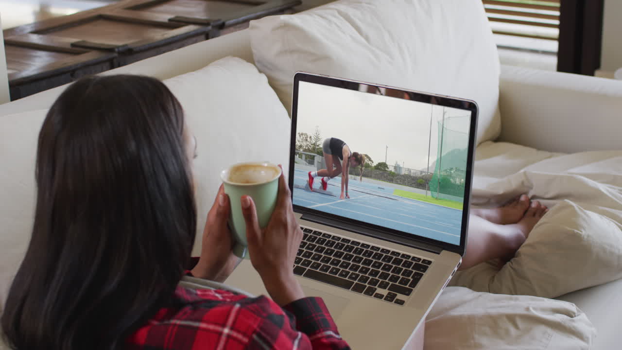 Composite of woman sitting at home holding coffee watching athletics running event on laptop